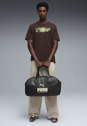 Mand iført brun Puma T-shirt og beige bukser, der holder en sort Puma sportstaske med teksten "Macht's mit Qualität" på grå baggrund.
