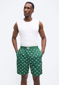 Homme portant un débardeur blanc et un short vert avec de petits logos crocodile blancs, debout les mains dans les poches.
