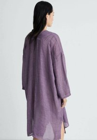 Violetti pellava-mekko, jossa on pitkät hihat, väljät leikkaukset ja sivuhalkiot. Kankaassa on teksturoitu pinta, ja se ulottuu reiden puoliväliin.