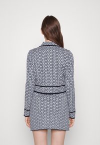 Kobieta ubrana w długą spódnicę blazerową z wzorem, w granatowy geometryczny print i granatowy wykończeniem, stojąca tyłem na jednolitym tle.