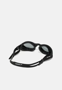 Lunettes de natation noires avec verres teintés, joints en silicone souple, sangle ajustable, et logo Speedo sur la sangle. Design léger et aérodynamique.