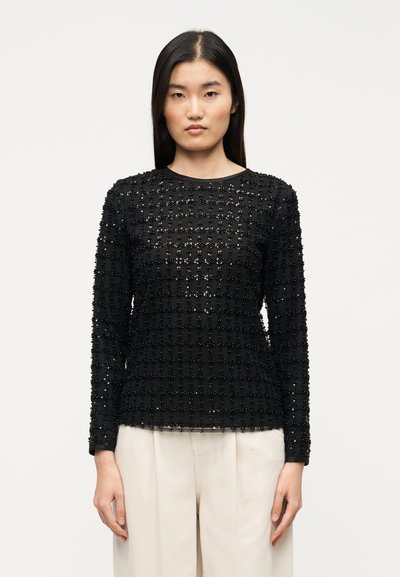 Schwarzes Langarmshirt mit strukturierter Stoffqualität und einem Raster aus Pailletten, rundem Ausschnitt und figurbetonter Silhouette. Kombiniert mit hellen Hosen.
