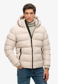 Chaqueta acolchada beige con capucha, cremallera negra y bolsillos laterales. Presenta puños acanalados y una textura suave y acolchada. Lleva puesta sobre un suéter de punto.