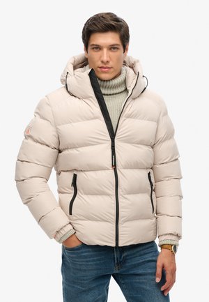 Chaqueta acolchada beige con capucha, cremallera negra y bolsillos laterales. Presenta puños acanalados y una textura suave y acolchada. Lleva puesta sobre un suéter de punto.