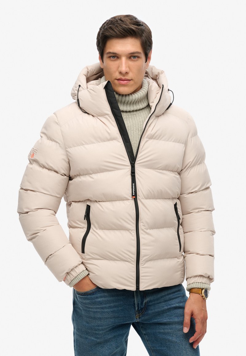 Chaqueta acolchada beige con capucha, cremallera negra y bolsillos laterales. Presenta puños acanalados y una textura suave y acolchada. Lleva puesta sobre un suéter de punto.