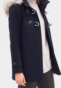 Navy blue winter coat with fur-trimmed hood, toggle closures, cuff button detail, worn over light gray sweater and dark pants.