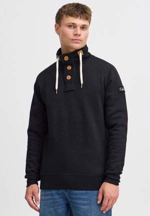 Czarny sweatshirt z wysokim kołnierzem, czterema drewnianymi guzikami i białymi sznurkami. Posiada naszywkę z logo na lewym rękawie.