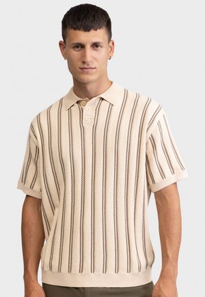 Νεαρός άνδρας με κοντά μαλλιά που φοράει μπεζ πλεκτό πόλο με κοντά μανίκια και κάθετες καφέ ρίγες, στέκεται μπροστά σε απλό φόντο.