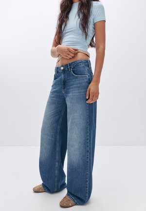 Wijduitlopende blauwe denim jeans met een vervaagde was, met voorzakken en een knoopsluiting. Gecombineerd met een lichtblauw aansluitend topje.