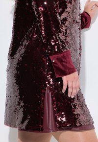 Vestido de lentejuelas burdeos con una textura brillante, que presenta una manga de terciopelo y una abertura lateral, destacando un diseño en capas y un acabado suave.