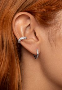 Pendientes de plata con un diseño curvado y piedras transparentes incrustadas en uno, y una forma afilada y angular en el otro, sobre fondo de cabello rojo.
