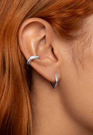 Pendientes de plata con un diseño curvado y piedras transparentes incrustadas en uno, y una forma afilada y angular en el otro, sobre fondo de cabello rojo.