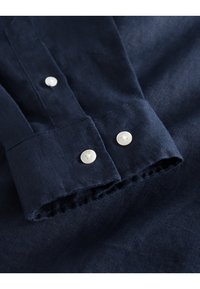 Laivastonsininen pellavapaita, jossa valkoiset napit. Paidassa on napillinen kaulus ja hieman teksturoitu kangas. Hihansuu näkyy, ja siinä on rispaantunut reuna.