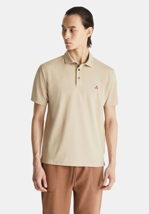Uomo che indossa una polo beige a maniche corte con logo rosso, abbinata a pantaloni marroni con coulisse, guarda verso sinistra su sfondo bianco.