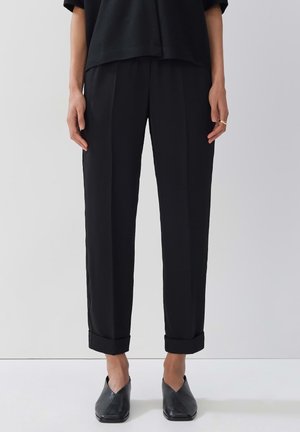 Schwarze, schmal zulaufende Hose mit glatter Textur, ausgestattet mit einem umgeschlagenen Saum und zwei vorderen Falten. Kombiniert mit schwarzen Slippern.