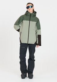 Ski-jack in lichtgroen en donkergroen met een capuchon, volledige rits en zijzakken; gecombineerd met zwarte skibroek en ski-botten.
