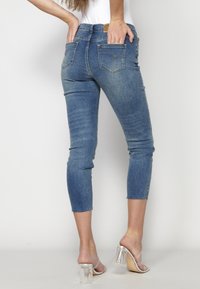 Jeans ajustados de color azul oscuro con un dobladillo desgastado, con dos bolsillos traseros y detalles de costura sutiles, combinados con zapatos de tacón transparente.