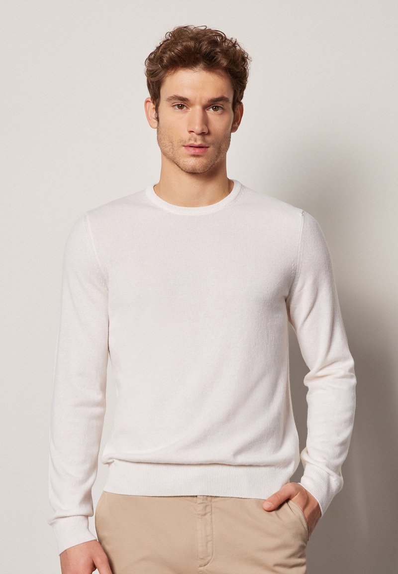 Weißer Pullover mit langen Ärmeln aus weichem Strickstoff, mit Rundhalsausschnitt und gerippten Bündchen. Getragen mit hellbeigen Hosen.