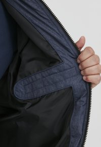 Mornarsko modra jakna s mehkim oblačilom, ki ima kontrastno temno notranjo plast, vidno zadrgo in oblikovano obris notranjega žepa.