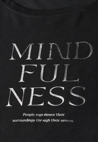 Černé bavlněné tričko s tučným stříbrným nápisem, který zní "MINDFULNESS", a menším textem uvádějícím "Lidé vnímají své okolí prostřednictvím svých smyslů."
