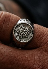 Sidabro žiedas su pakeltu dizainu, vaizduojančiu stilizuotą figūrą, geometrinius motyvus ir užrašą "EAST" ant juostos. Tekstūruota paviršiaus detalė.