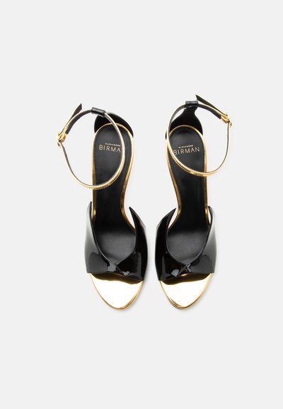 Μαύρα και χρυσά ανοιχτά σανδάλια Alexandre Birman με λουριά στον αστράγαλο, σε θέαση από ψηλά πάνω σε λευκό φόντο.