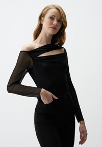 Schwarzer langärmeliger Bodysuit mit durchsichtigen Mesh-Ärmeln, einem gedrehten Ausschnitt und Aussparungen, kombiniert mit hoch taillierten schwarzen Jeans.