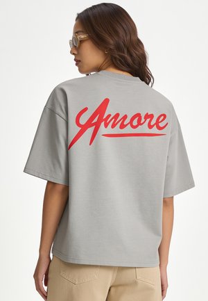 Vrouw met lang bruin haar draagt een grijs oversized T-shirt met rode tekst "Amore" op de rug en beige broek, kijkt naar links.