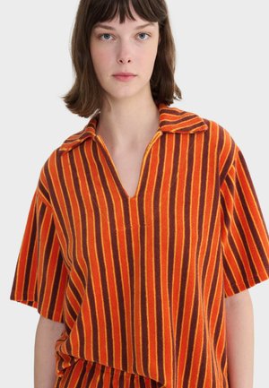 Mladá žena s krátkymi hnedými vlasmi, oblečená v voľnej košeli s golierom a krátkymi rukávmi s vertikálnymi oranžovými a červenými pruhmi.