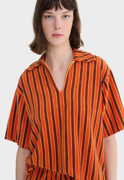 Jeune femme aux cheveux bruns courts portant une chemise ample à col et manches courtes avec des rayures verticales orange et rouges.