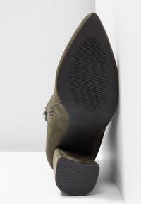 Botte de cheville en daim vert olive avec un bout pointu, semelle en caoutchouc noir texturé et détail de fermeture éclair sur le côté. Talon plat et design angulaire.