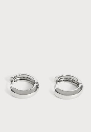 Paire de petites boucles d’oreilles créoles argentées brillantes avec surfaces lisses et arrondies sur fond blanc uni.