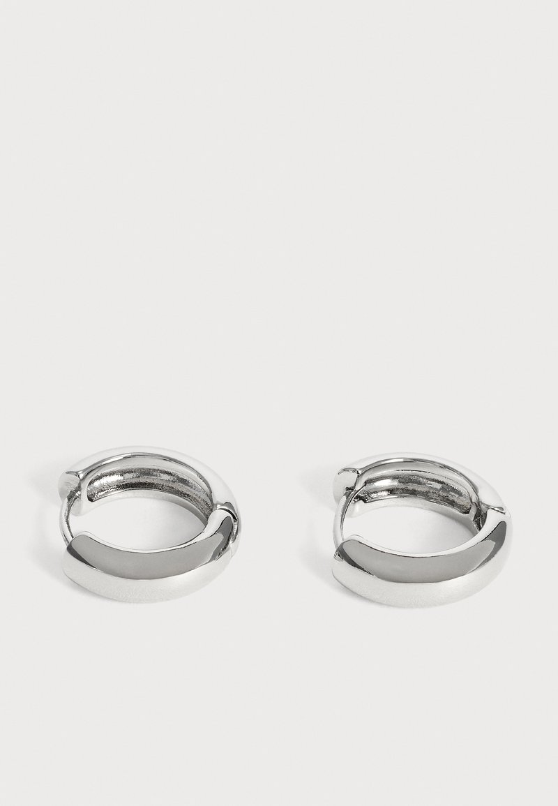 Paire de petites boucles d’oreilles créoles argentées brillantes avec surfaces lisses et arrondies sur fond blanc uni.