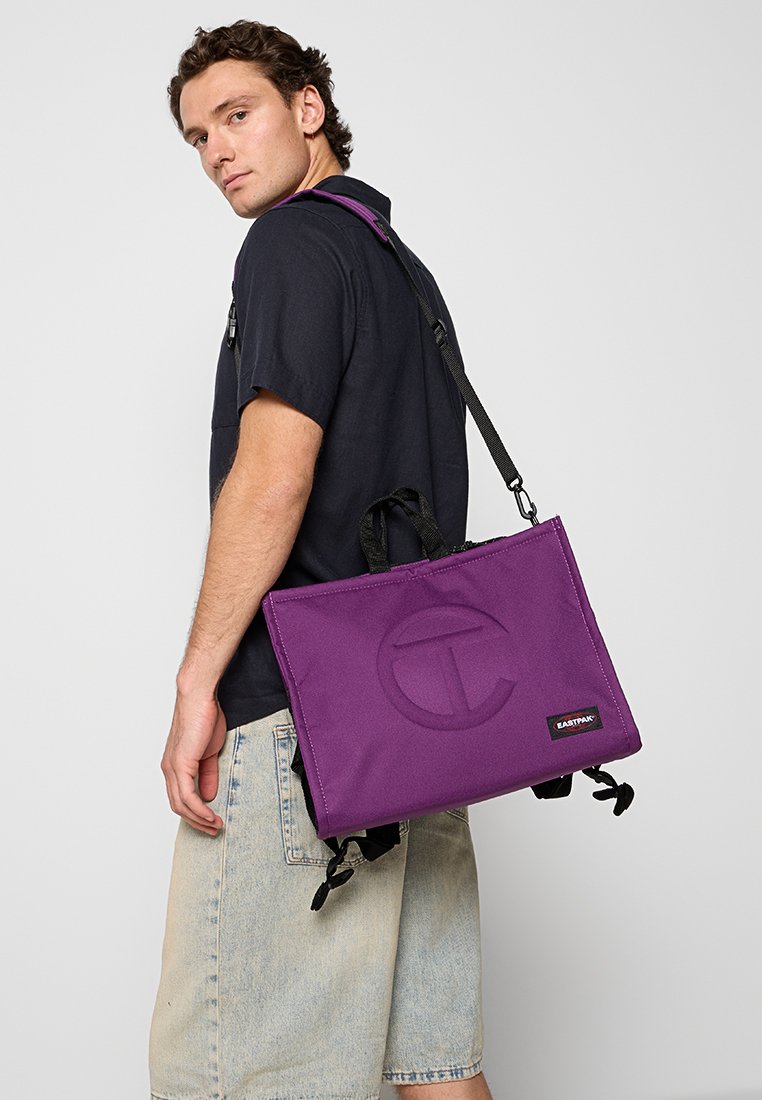 Borsa a tracolla in tessuto viola con motivo a logo, manici corti e una lunga tracolla regolabile, con dettagli neri e una texture liscia.