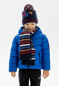 Veste doudoune bleue, bonnet en tricot marine avec pompon et texte « HEY », écharpe à rayures multicolores, pantalon noir. Texture lisse et design décontracté.