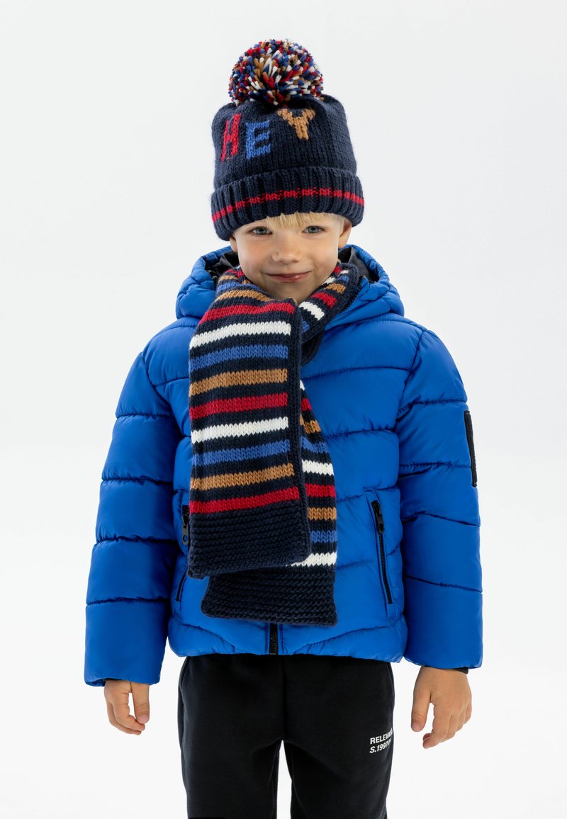 Veste doudoune bleue, bonnet en tricot marine avec pompon et texte « HEY », écharpe à rayures multicolores, pantalon noir. Texture lisse et design décontracté.