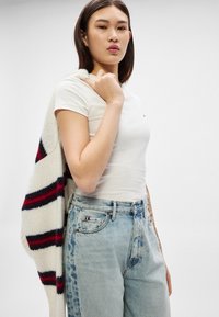 Jauna sieviete baltā cieši piegulošā T-kreklā un gaišzilās džinsās, turot baltu adītu džemperi ar sarkani melnām svītrām pār plecu.