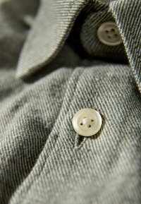 Graues Hemd mit Kragen aus strukturiertem Stoff, mit einem hellfarbenen Vierlochknopf und sichtbaren Nahtdetails.