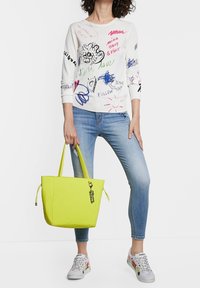 Limonengrüne Tasche aus Textil mit geprägter Struktur, breiten Seiten und kurzen Griffen. Ausgestattet mit einem schwarzen Charm mit der Aufschrift "Jetzt." Kombiniert mit einem weißen Grafikoberteil und Jeans.