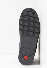 Semelle d'une botte Hunter noire montrant des motifs d'adhérence texturés, taille UK 4, fabriquée en Chine, avec un logo circulaire rouge près du talon.