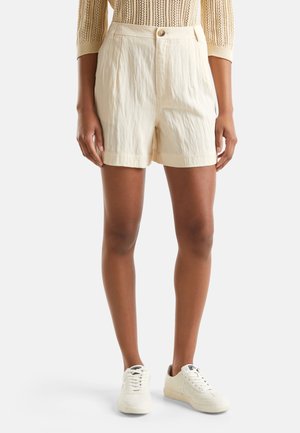 Personne portant un short beige texturé, un haut en maille beige à manches longues et des baskets blanches, debout devant un fond uni.