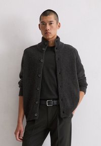 Cardigan en tricot gris foncé avec un col haut, cinq boutons à l'avant, des poignets côtelés et une finition texturée, associé à une chemise noire.