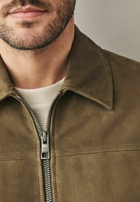 Bruine suede jack met een ritssluiting aan de voorkant, puntige kraag en een geribbeld wit onderhemd zichtbaar eronder. Glad textuur en een strak ontwerp.