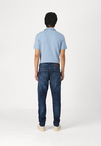 Lichtblauw polohemd met korte mouwen gecombineerd met donkerblauwe denim jeans. De jeans hebben zichtbare stiksels, achterzakken en een leren patch op de tailleband.