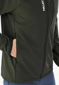 Veste softshell vert foncé avec une fermeture éclair à l'avant, des poignets élastiques et une poche zippée sur le côté pour un rangement sécurisé.
