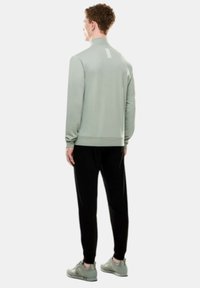 Giacca sportiva verde chiaro, aderente, con una texture liscia, colletto alto, caratterizzata da accenti bianchi sulla schiena, abbinata a pantaloni neri a gamba affusolata e sneaker grigie.