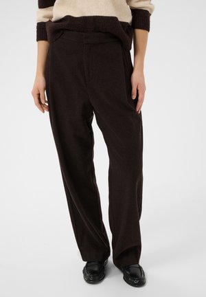 Personne portant un pantalon ample marron foncé, des mocassins noirs et un pull beige et marron rayé, debout devant un fond uni.