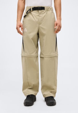 Beige Herren-Convertible-Hosen mit schwarzen Akzenten, verstellbarem Bund und Reißverschluss an den Knien, getragen mit schwarzen Schuhen und weißem Oberteil.