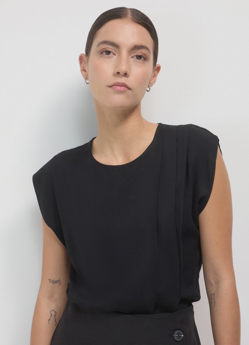 Blouse noire en tissu léger et doux, avec des manches cape surdimensionnées et des plis verticaux d'un côté. Encolure haute avec des lignes épurées.
