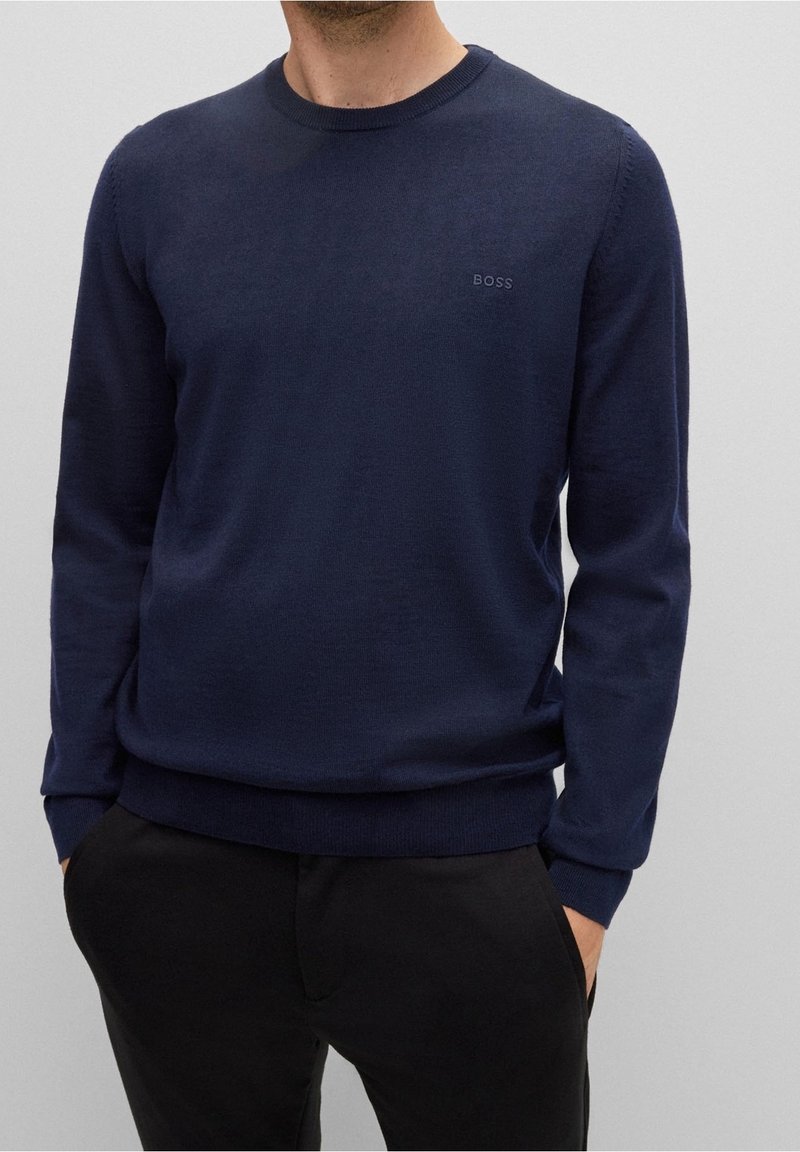 Homme portant un pull à col rond à manches longues bleu marine avec le logo "BOSS" sur le côté gauche de la poitrine et un pantalon noir, les mains dans les poches, sur un fond uni.